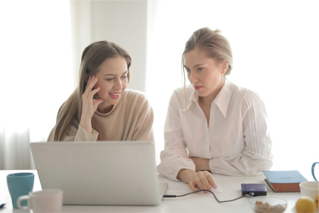 Zwei Frauen sitzen an einem Tisch und schauen in einen Laptop. Sie grübeln. 