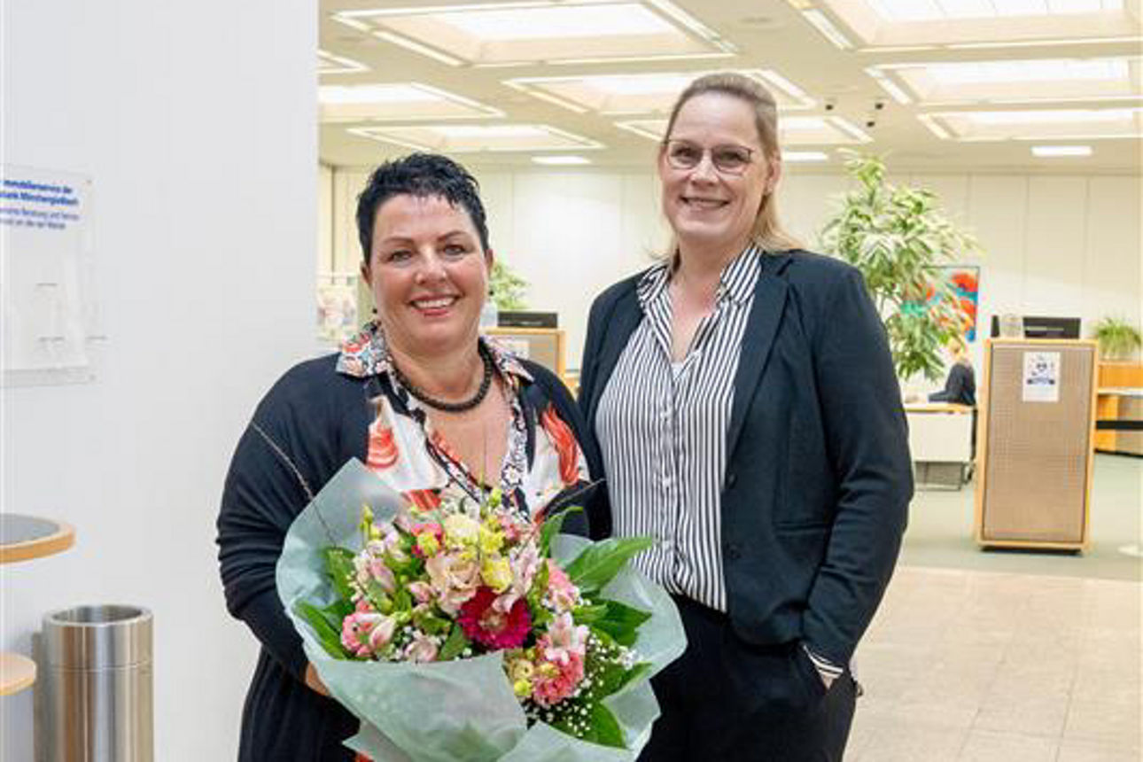 Zwei Frauen stehen nebeneinander und lächeln in die Kamera während eine der beiden einen Blumenstrauß hält