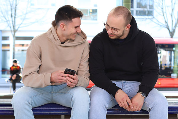 Zwei Kollegen sitzen an der Bahnhaltestelle auf einer Bank und schauen gemeinsam in ein Handy