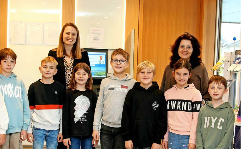 Eine Gruppe von Kindern steht vor einer Frau in einer Bluse mit Leopardenmuster und einer Frau in einem braunen Pullover.