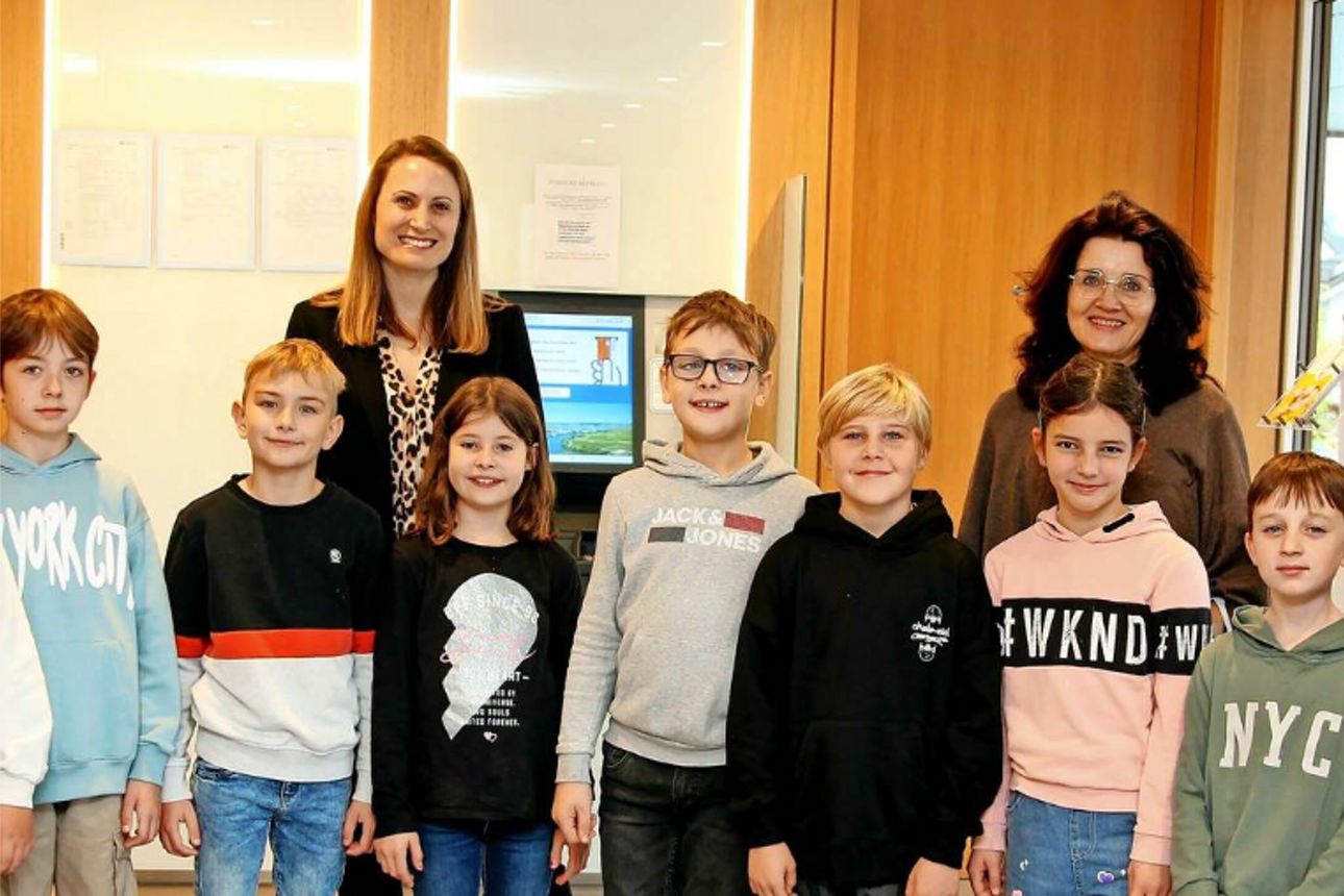Eine Gruppe von Kindern steht vor einer Frau in einer Bluse mit Leopardenmuster und einer Frau in einem braunen Pullover.