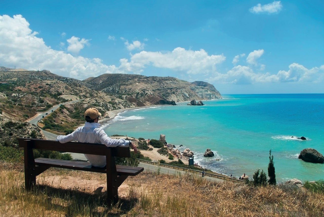 Ein Mann sitzt auf einer Bank auf einem Aussichtspunkt und schaut auf das blau / türkis strahlende Meer 