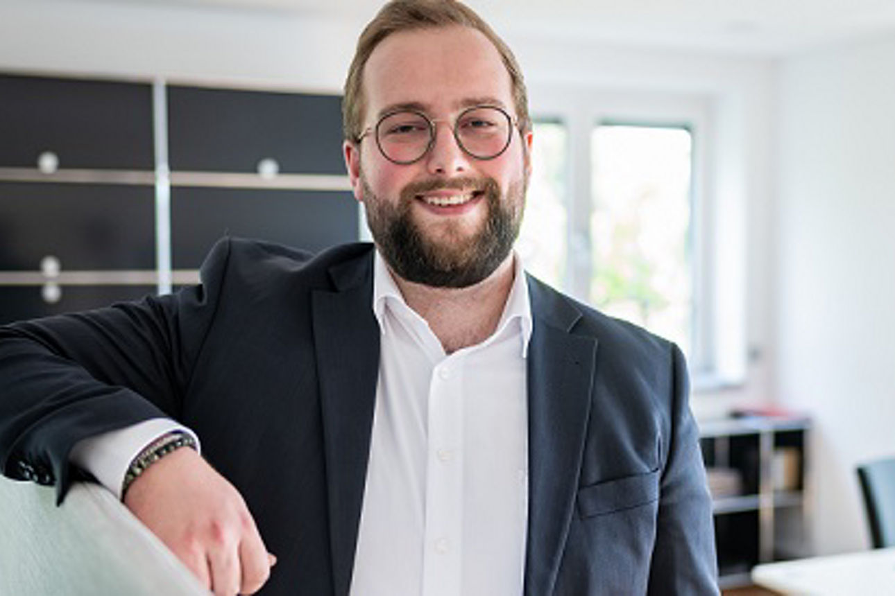 Junger Mann lehnt an eine Stellwand und lächelt dabei