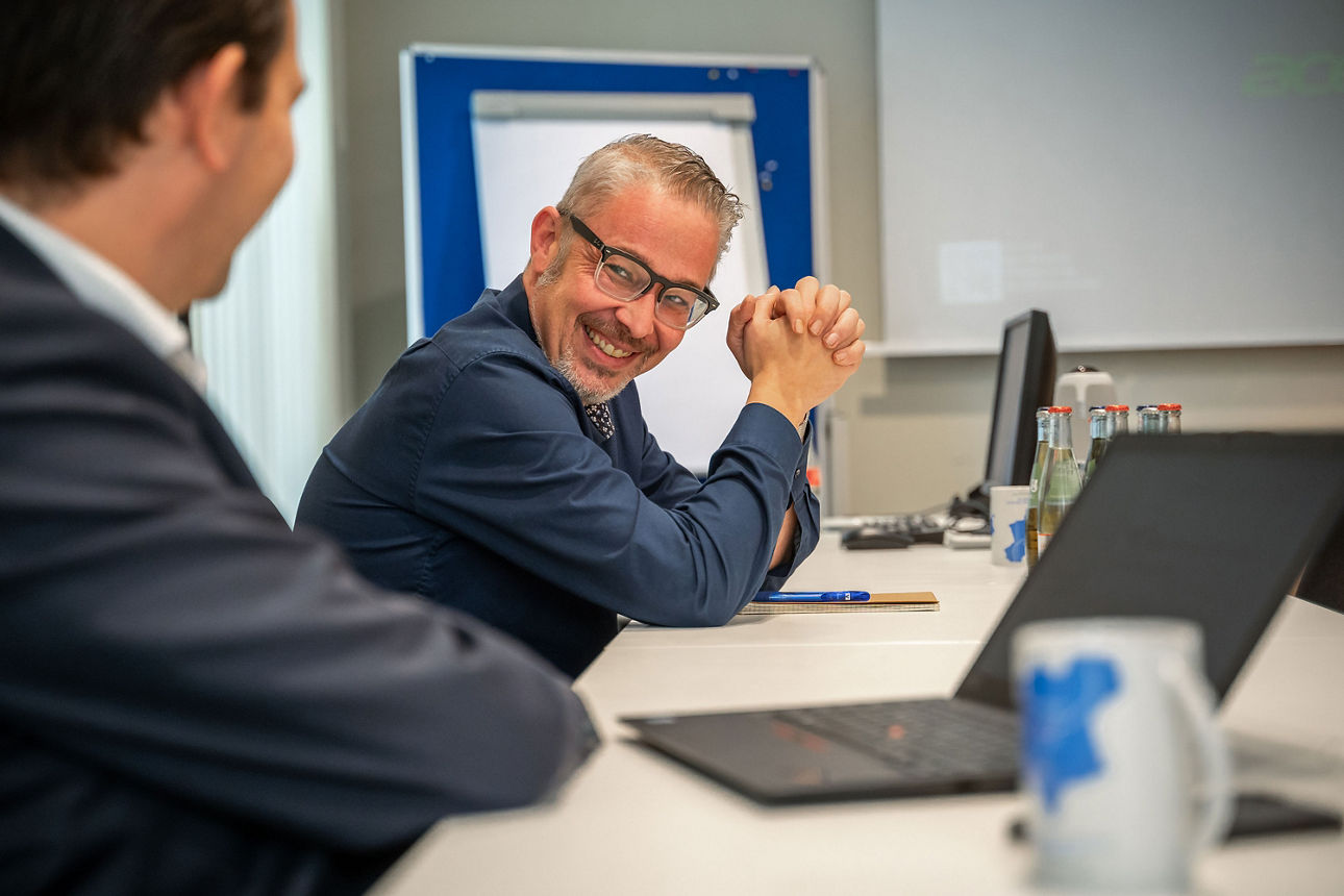 Zwei Männer sitzen lächelnd am Schreibtisch in der Bank