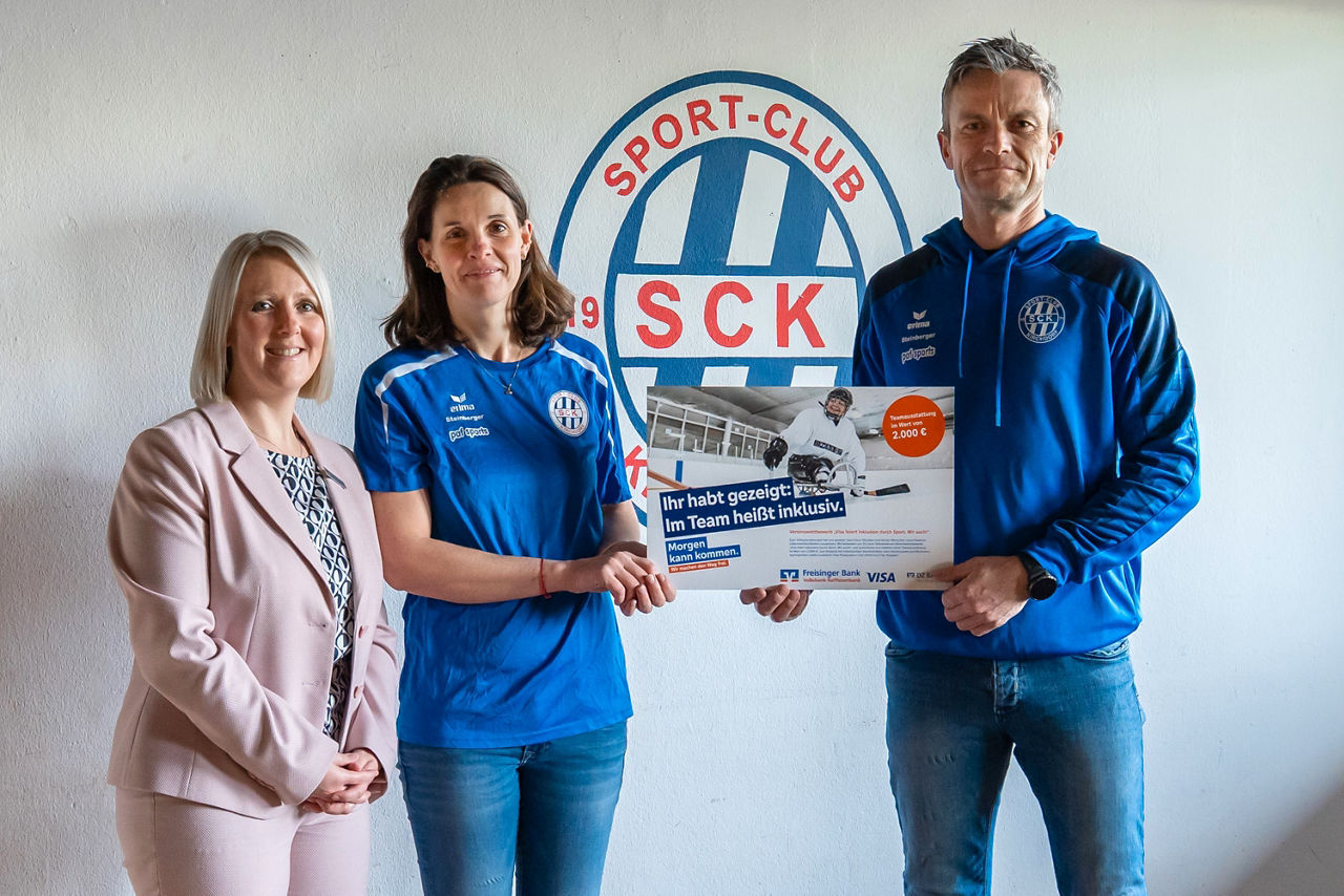 Zwei Frauen und ein Mann stehen vor einer weißen Wand. Auf der Wand ist das Logo vom SC KirchdorfDer Mann und die Frau in der Mitte halten ein kleines Plakat in der Hand , sie tragen Kleidung des Sportvereins. Di e Frau links trägt einen rosé-farbenen Hosenanzug