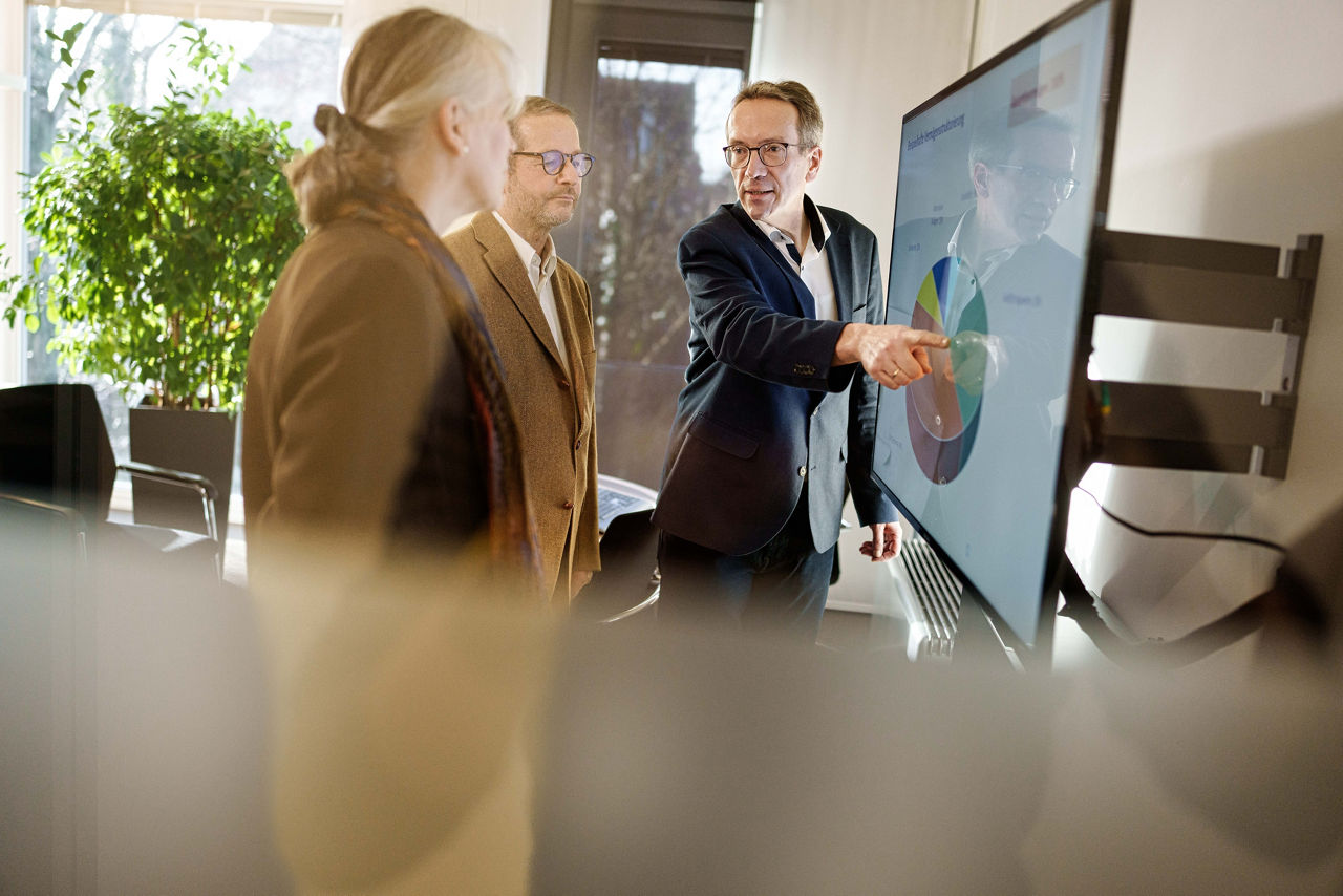 Vermögenskundenbetreuer steht mit zwei Kunden vor einem Monitor und zeigt darauf