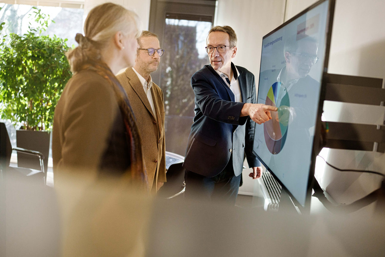 Vermögenskundenbetreuer steht mit zwei Kunden vor einem Monitor und zeigt darauf