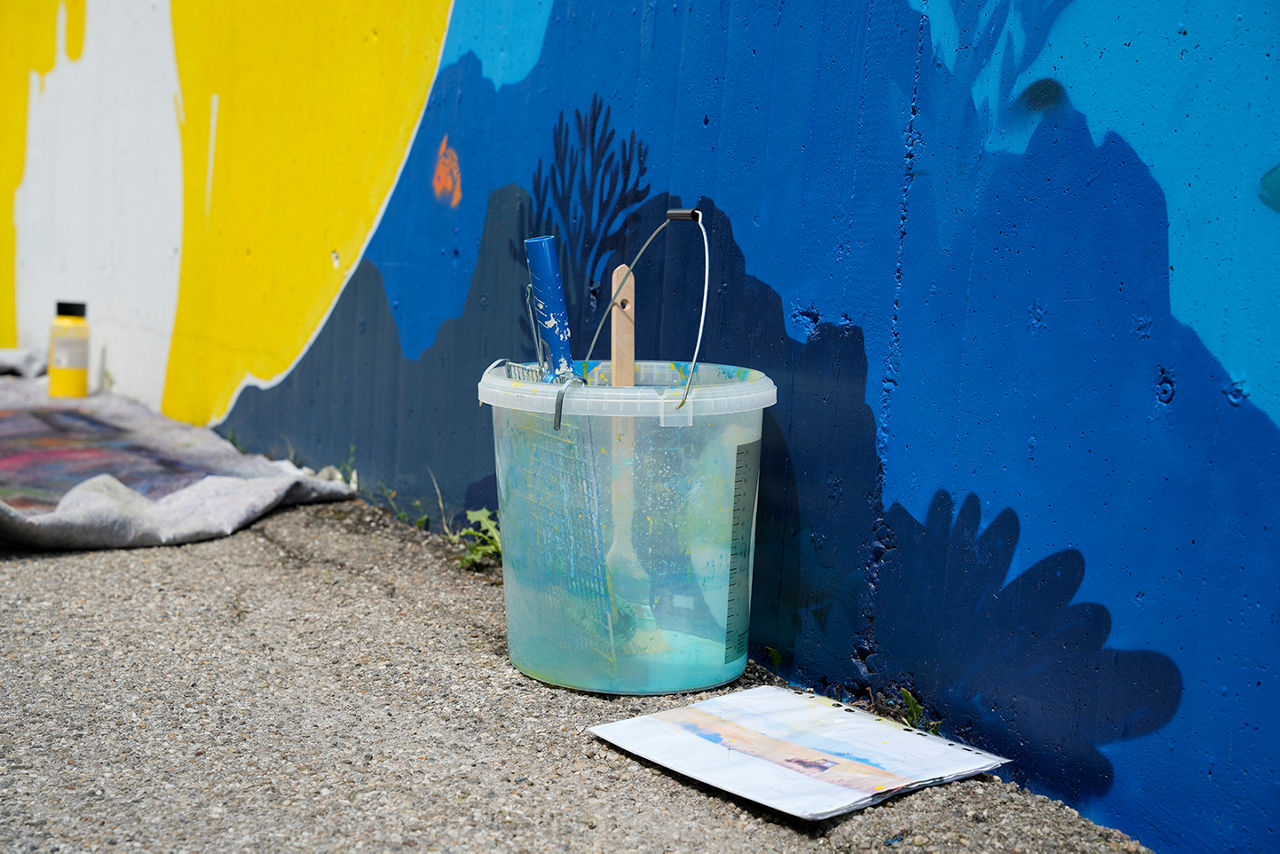 Farbeimer steht vor blau-gelber Wand, auf der ein Kunstwerk entsteht