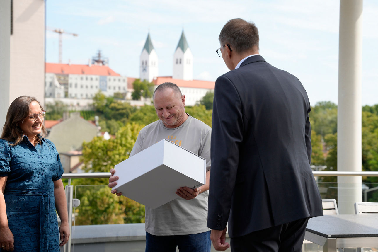 Ein Mann nimmt auf einer Terrasse ein großes weißes Geschenk entgegen, während zwei weitere Personen lächelnd danebenstehen. Im Hintergrund ist eine Stadtsilhouette von Freising mit dem Dom zu sehen