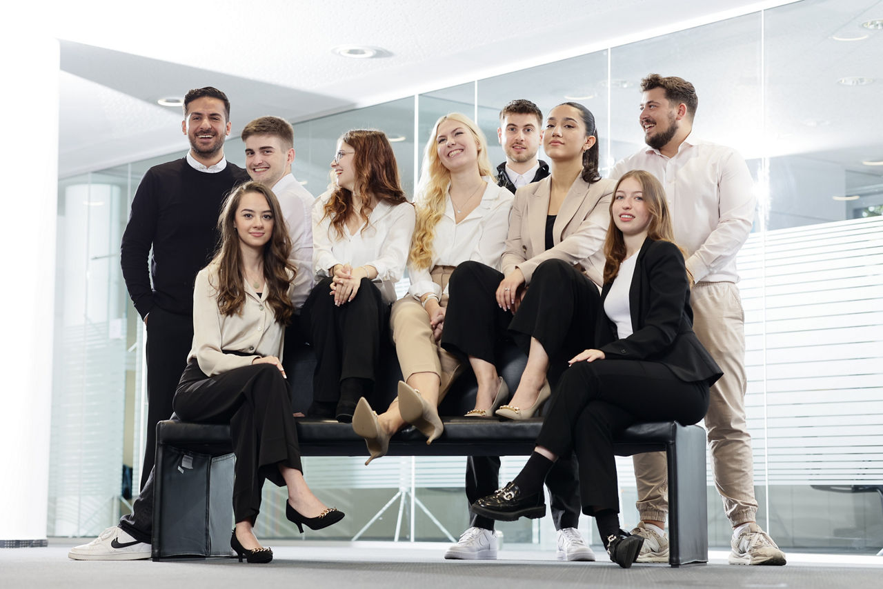 Eine Gruppe von neun jungen Frauen und Männern sitzt und steht um bzw. auf einer Bank in einem geschäftlichen Raum