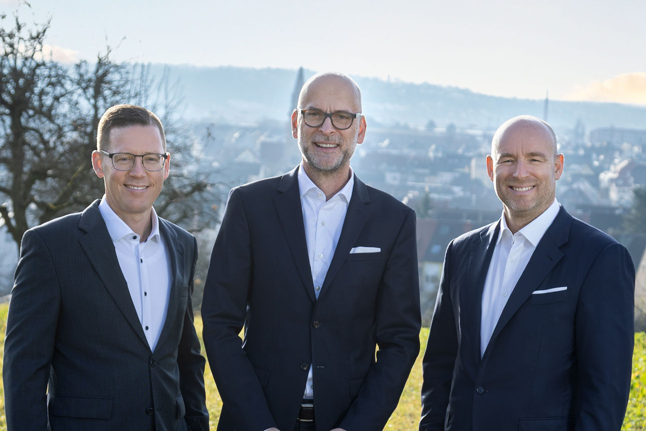 Drei Männer im Anzug stehen vor einer Baumwiese