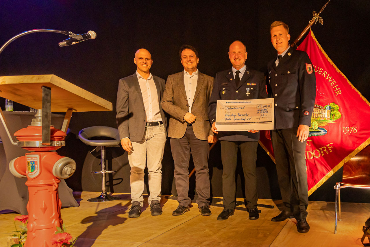 Vorstände der Feuerwehr und der Bank präsentieren einen Scheck in Höhe von 7.000 Euro auf einer Bühne