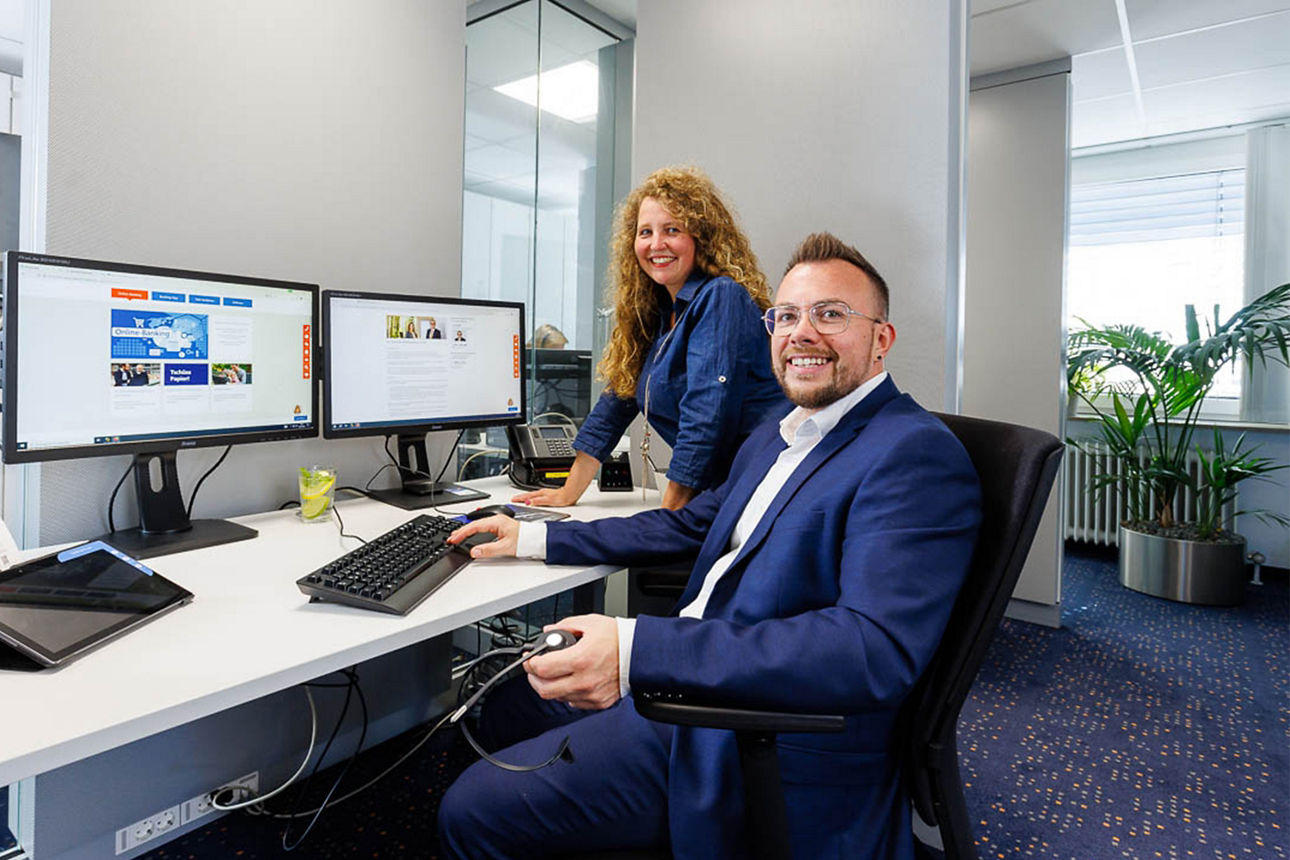 KundenDialogcenter - Frau mit roten Haaren steht am Schreibtisch hinter einem Mann mit Brille, beide Lachen freundlich.