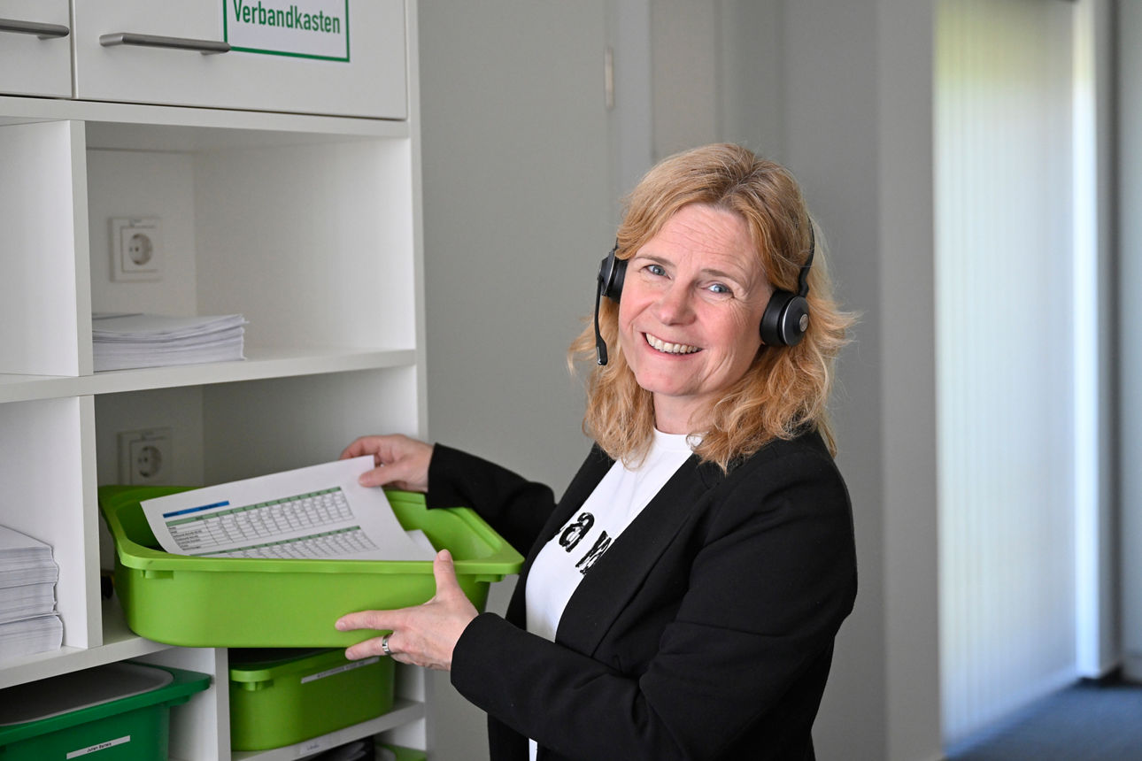 Eine Mitarbeiterin der Digitalfiliale steht mit einer grünen Kiste mit Unterlagen am Regal