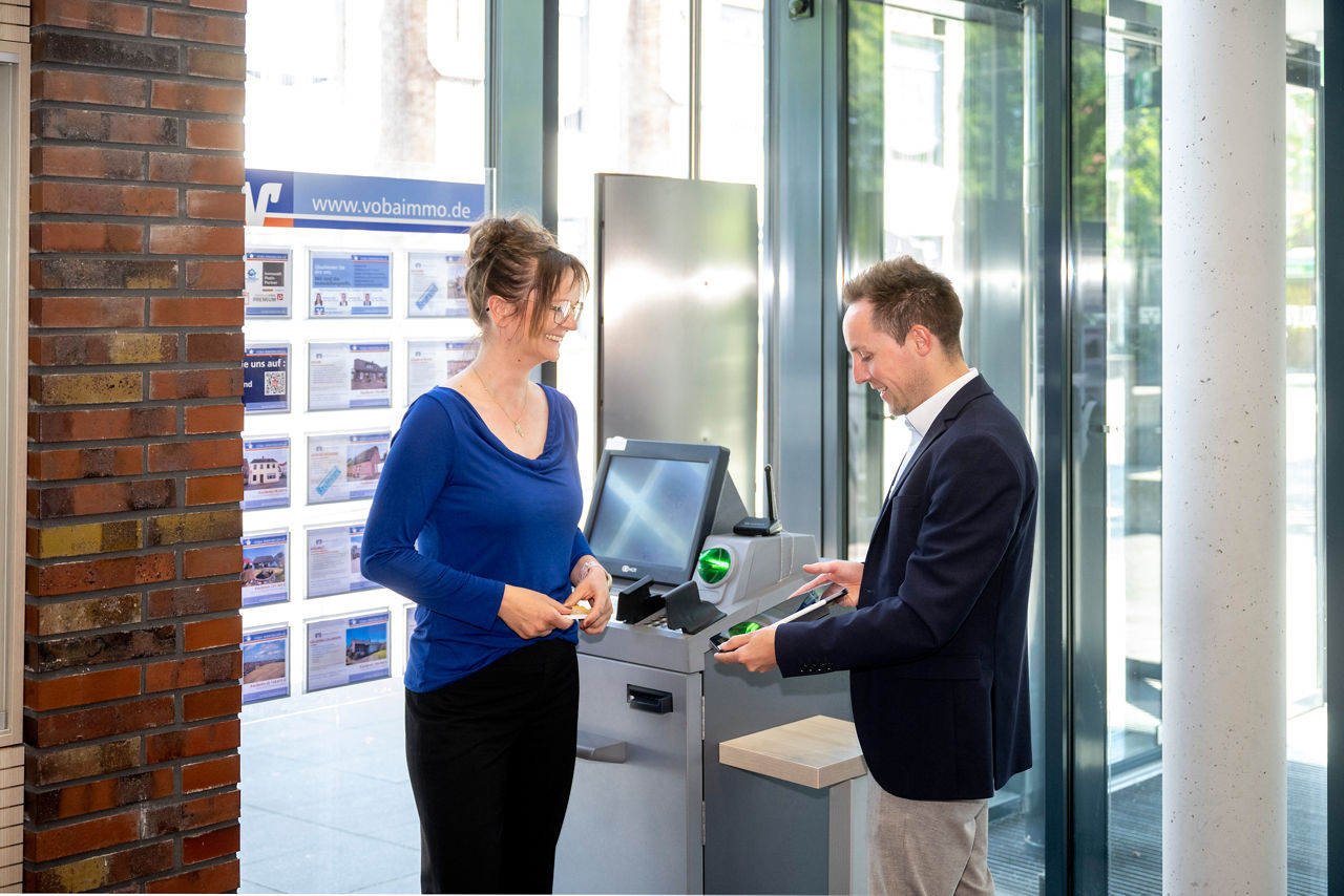 Ein Berater erklärt der Kundin etwas vor dem Überweisungsterminal