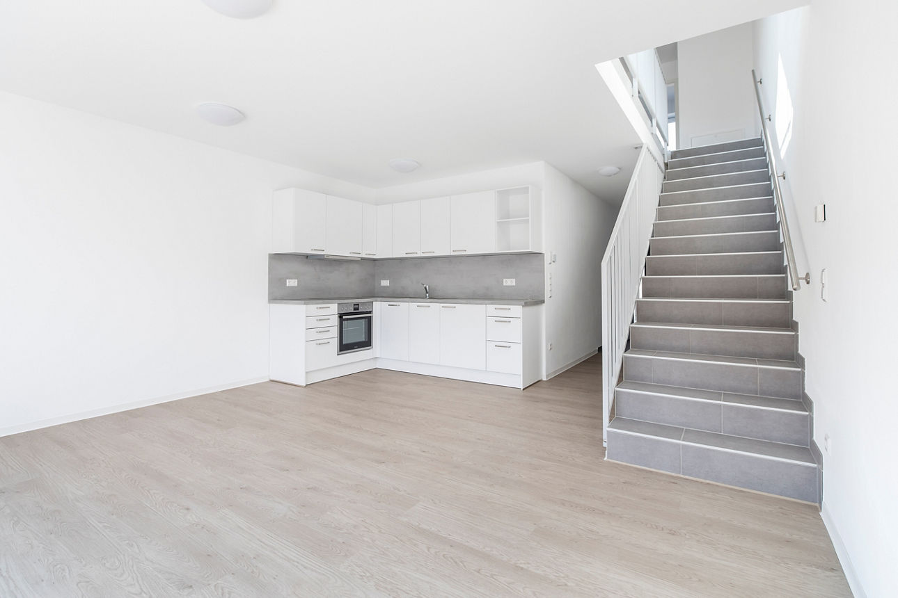 Darstellung einer weißen kleinen Küchenzeile sowie einem großzügigen Wohnbereich sowie einer Treppe ins Obergeschoss eines modernen, energieautarken Mehrfamilienhauses.