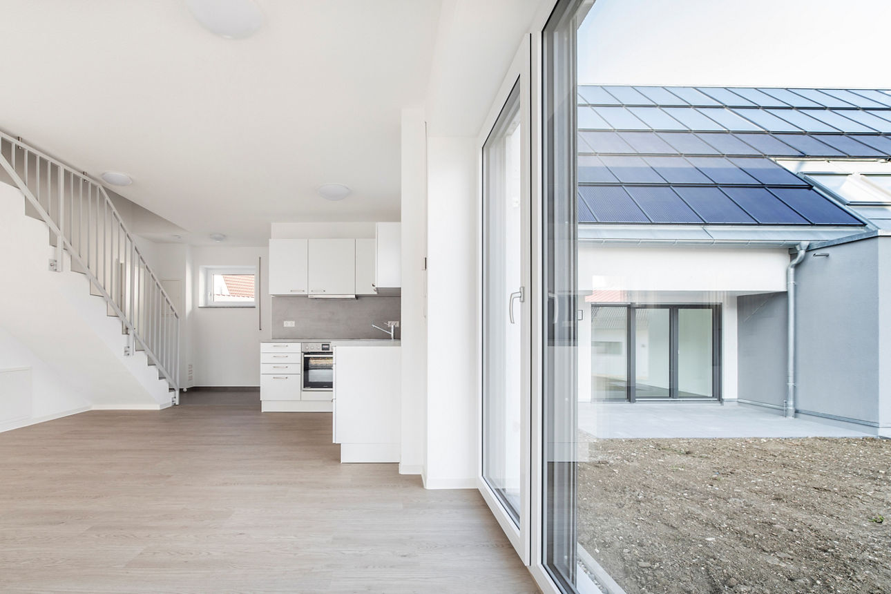 Darstellung einer kleinen, weißen Küchenzeile sowie eines großzügigen Wohnbereich in einem modernen, energieautarlen Haus. Auf der linken Seite ist eine Treppe ins OG zu sehen. Auf der rechten Seite ist eine große Fensterfront zu sehen mit Blick auf das benachbarte spiegelbildliche energieautarke Mehrfamilienhaus. 