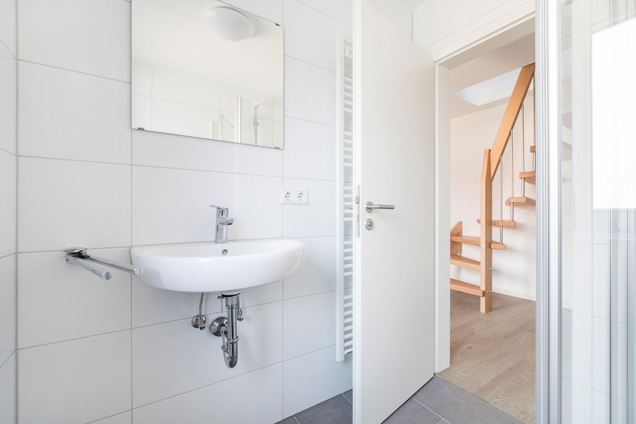 Darstellung der Toilette im EG eines modernen, energieautarken Mehrfamilienhauses. Zu sehen ist ein weißes Waschbecken sowie ein Spiegel darüber. Durch die geöffnete Tür ist die Holztreppe im bereich des Windfangs zu sehen, welche ins OG führt. 