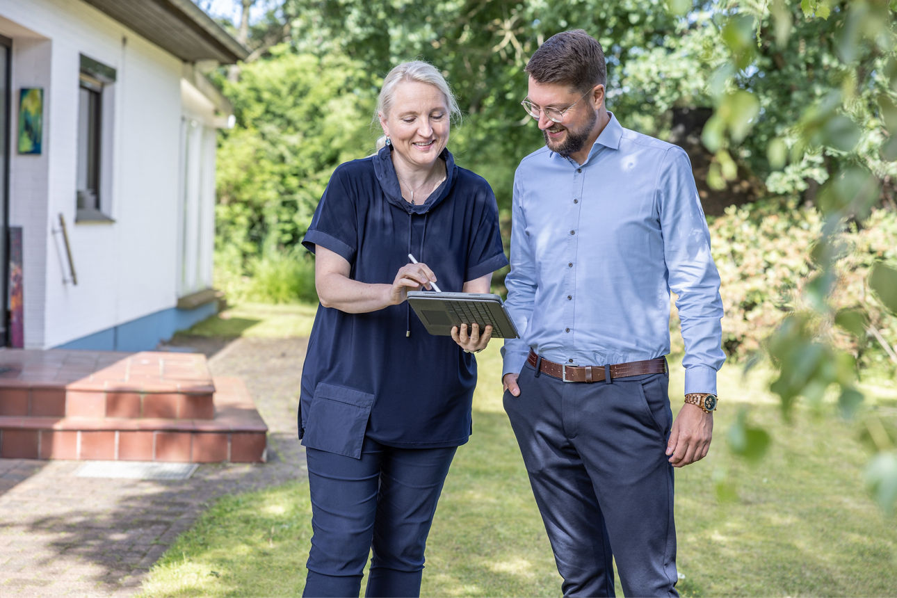 Zwei Personen stehen im Garten, eine hält ein Tablet und zeigt darauf, die andere schaut zu, 