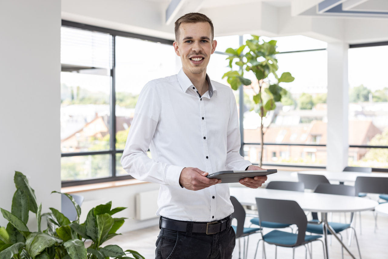 Mann in weißem Hemd und dunkler Hose hält Tablet in modernem Büro mit Pflanzen und Stühlen.