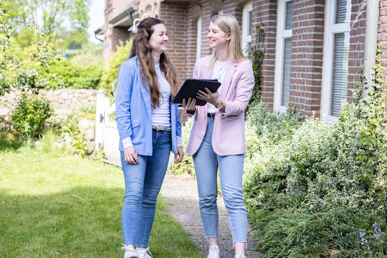Zwei Frauen stehen vor einem Backsteinhaus umgeben von Gartenpflanzen.