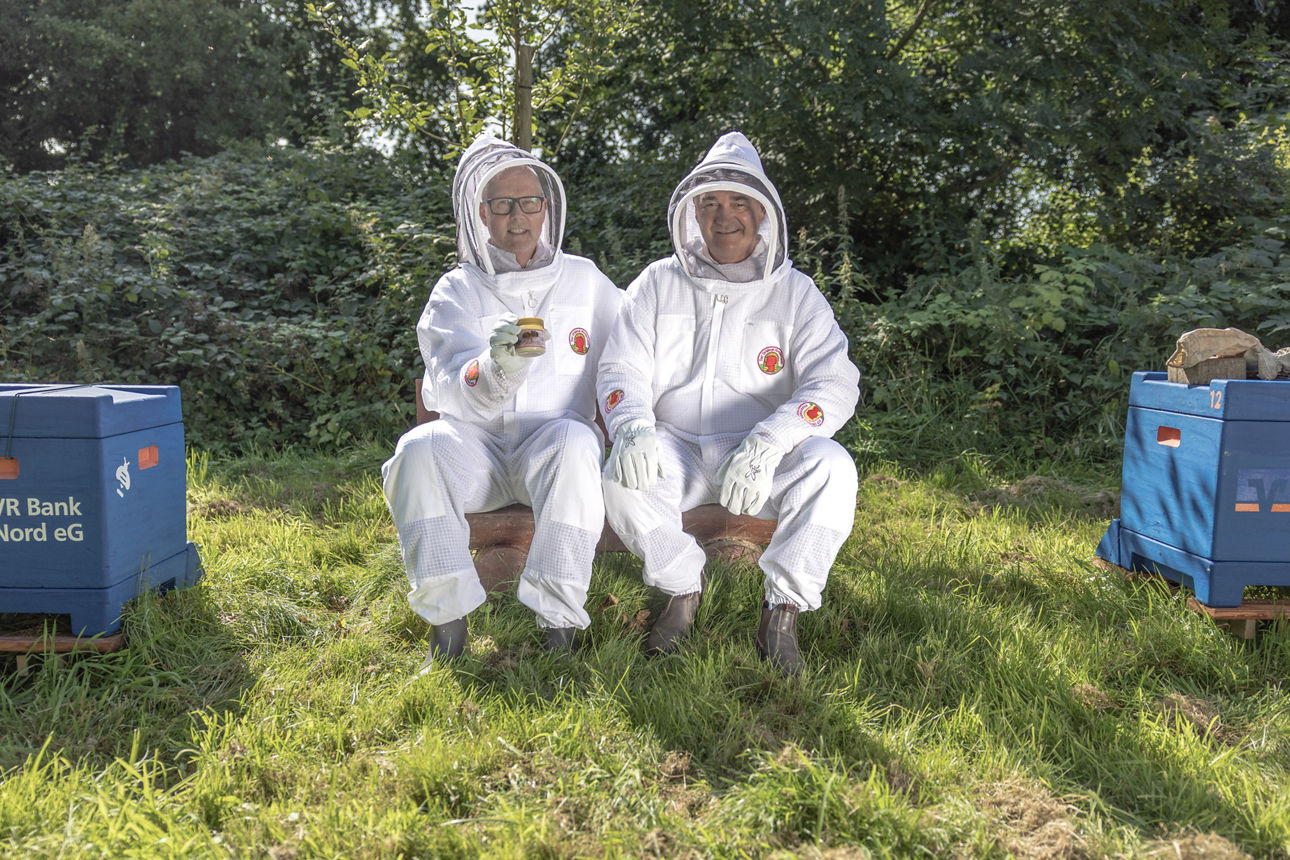 Zwei Männer in Imkerschutzanzügen sitzen auf einer Bank, einer der Männer hält ein Glas Honig in der Hand.