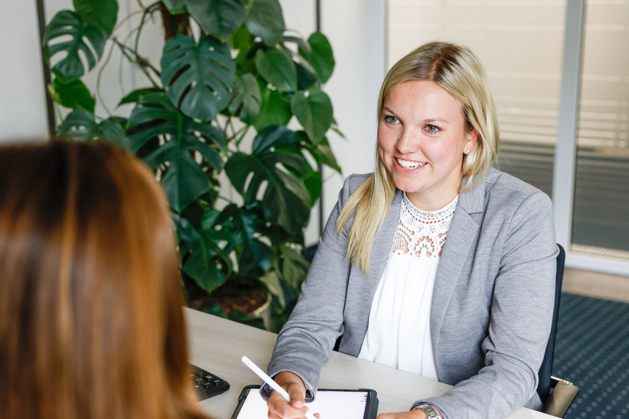 Zwei Frauen sitzen an einem Tisch, eine hält einen Stift und ein Tablet, im Hintergrund eine große grüne Pflanze.