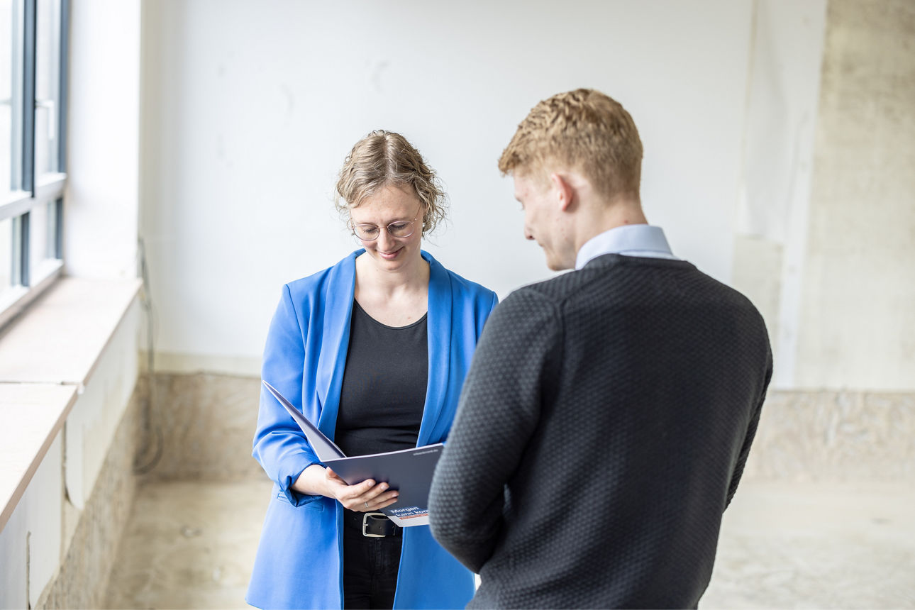 Zwei Personen stehen sich in einem hellen Raum mit großen Fenstern gegenüber, eine hält einen Laptop in der Hand
