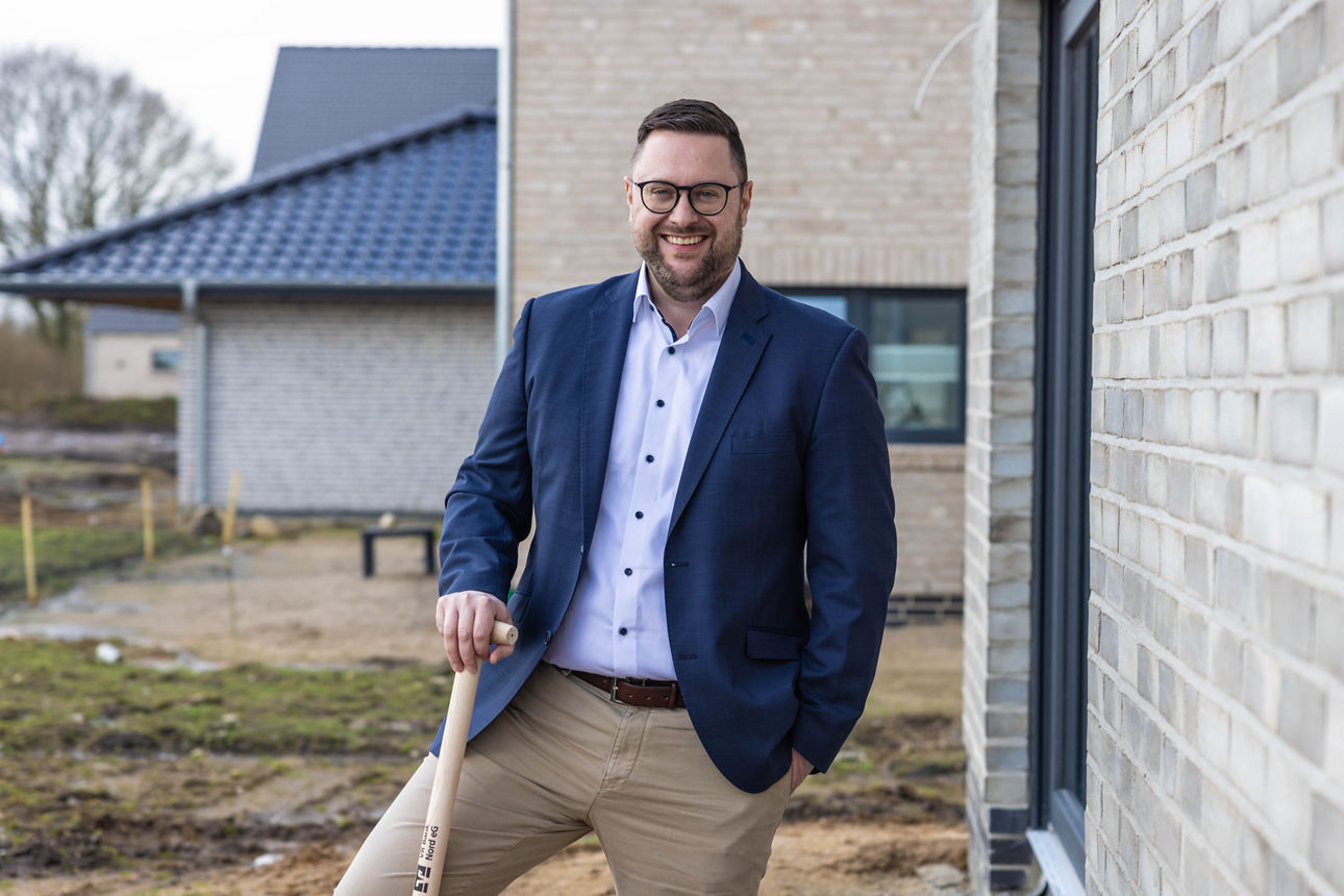 Ein Mann steht vor einem Neubau und stützt sich mit der rechten Hand auf einer Schaufel ab.