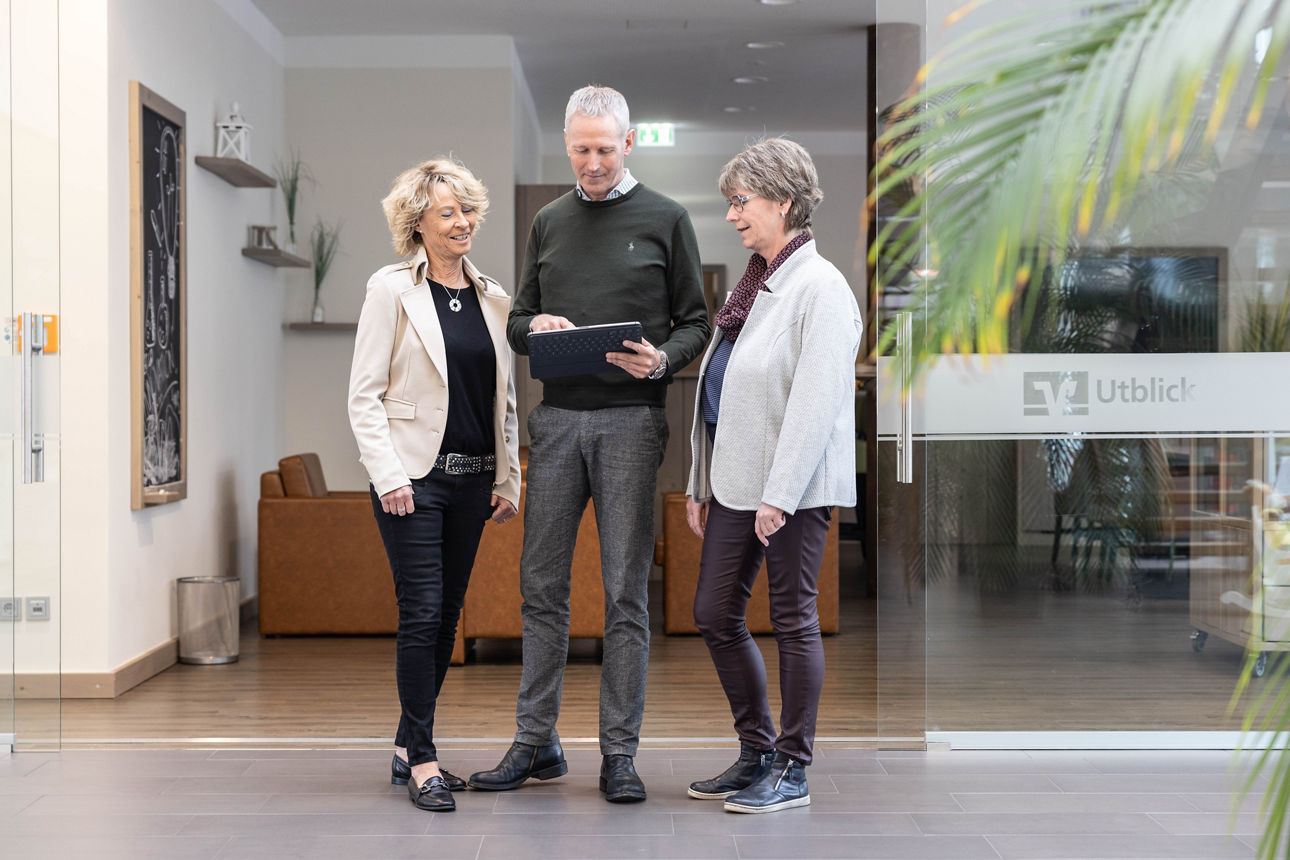 Drei Personen stehen in einem hellen Flur vor einer Glastür mit dem Schriftzug 'Utblick', eine Person hält ein Tablet, alle tragen Business-Kleidung.
