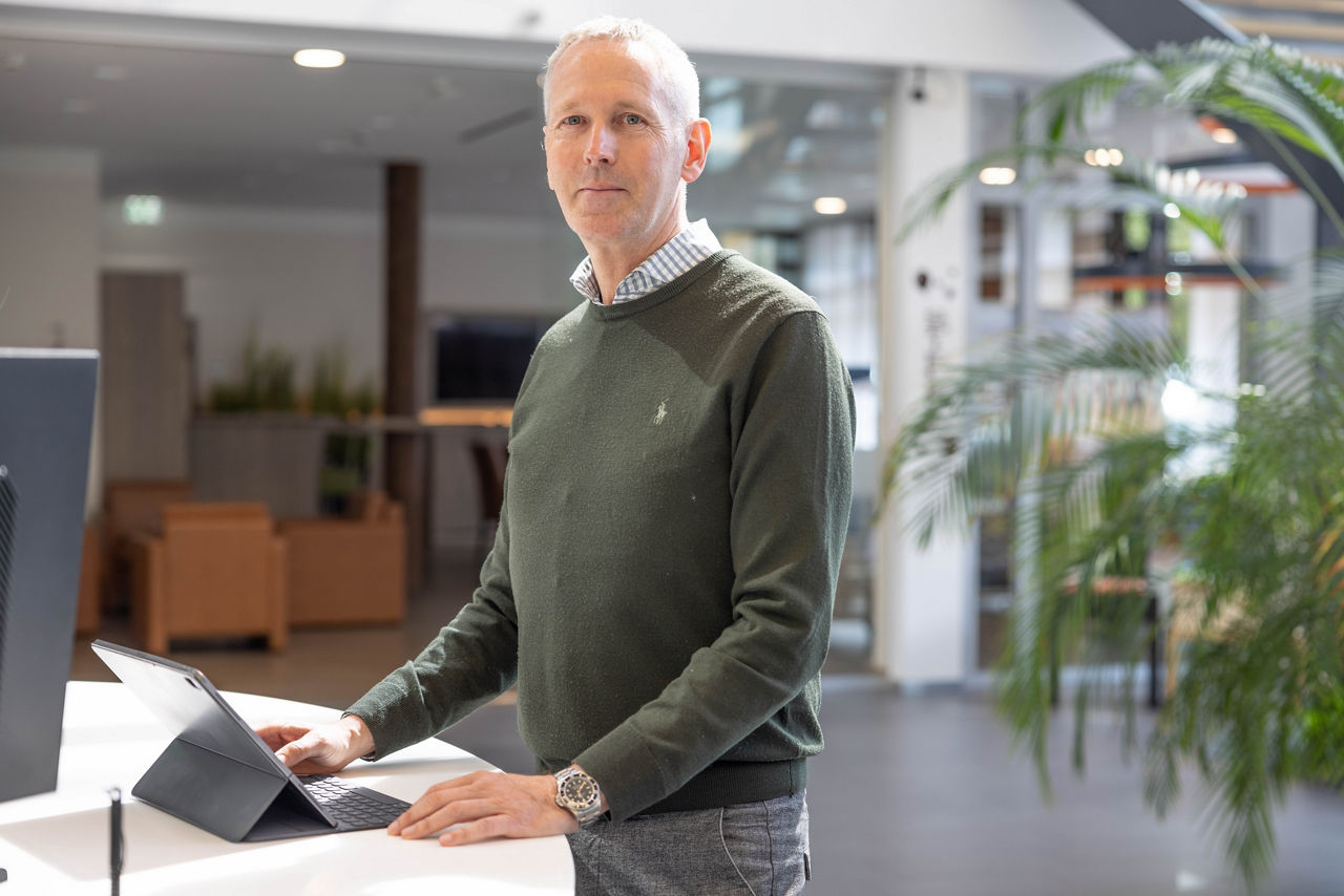 Person mit grünem Pullover und Hemd steht an einem weißen Tisch und arbeitet an einem Tablet mit Tastatur in einem modernen Büro.