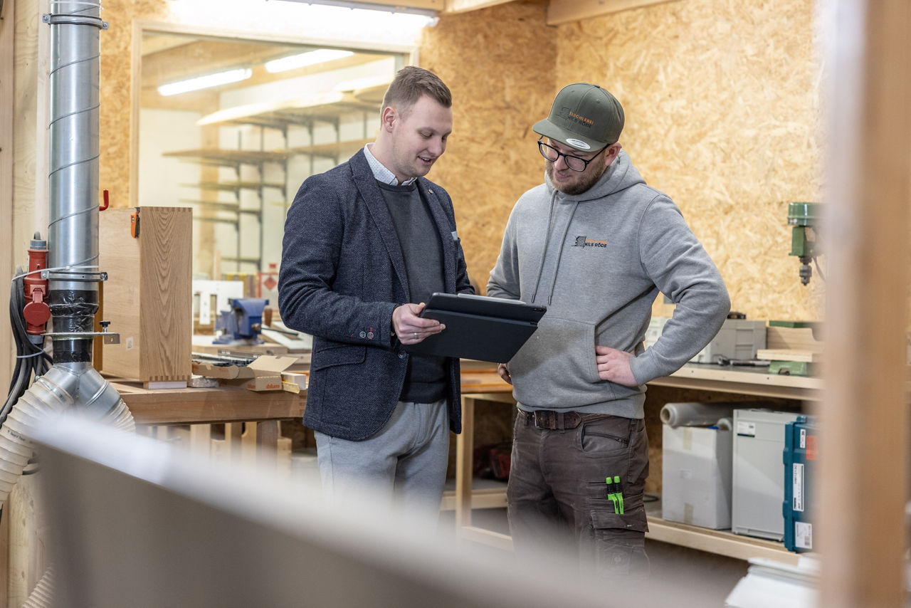 Firmenkundenbetreuer steht mit seinem Kunden in einer Werkstatt und schaut auf ein Tablet.