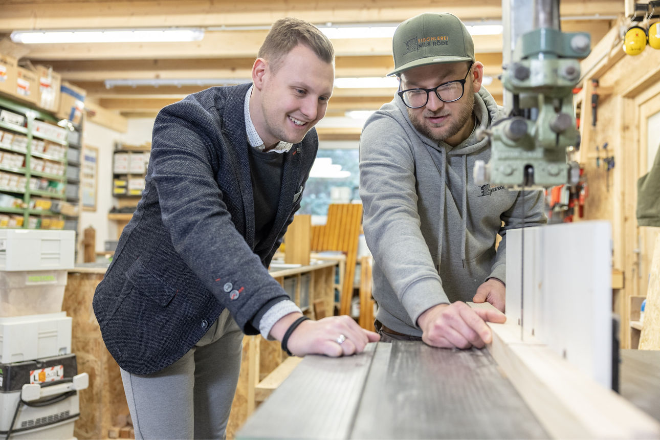Firmenkundenbetreuer steht mit seinem Kunden in einer Werkstatt und schaut beim Holz schneiden zu