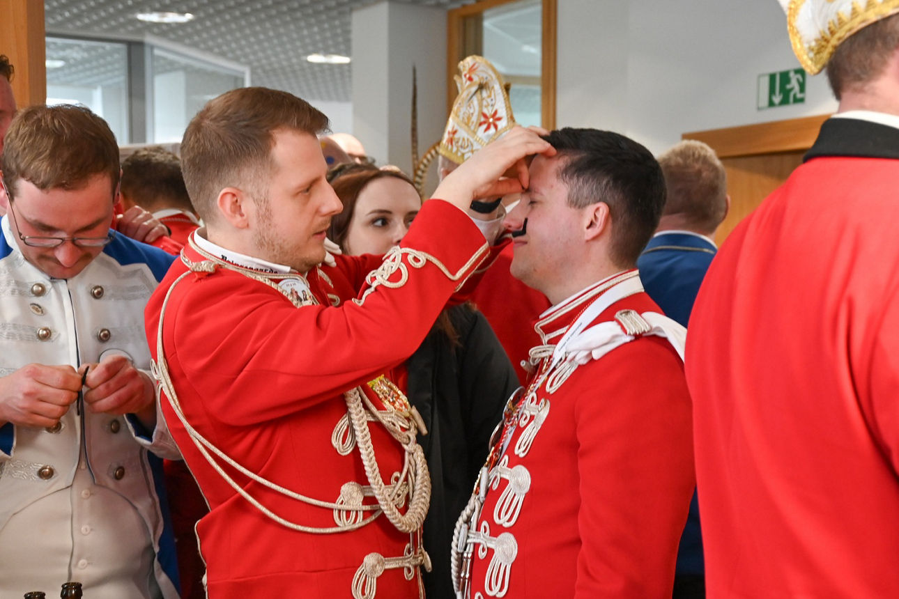 Ein Mann in rot-weißer Garde-Uniform klebt einem Vereinskameraden falsche Schnäuzer als Augenbrauen an