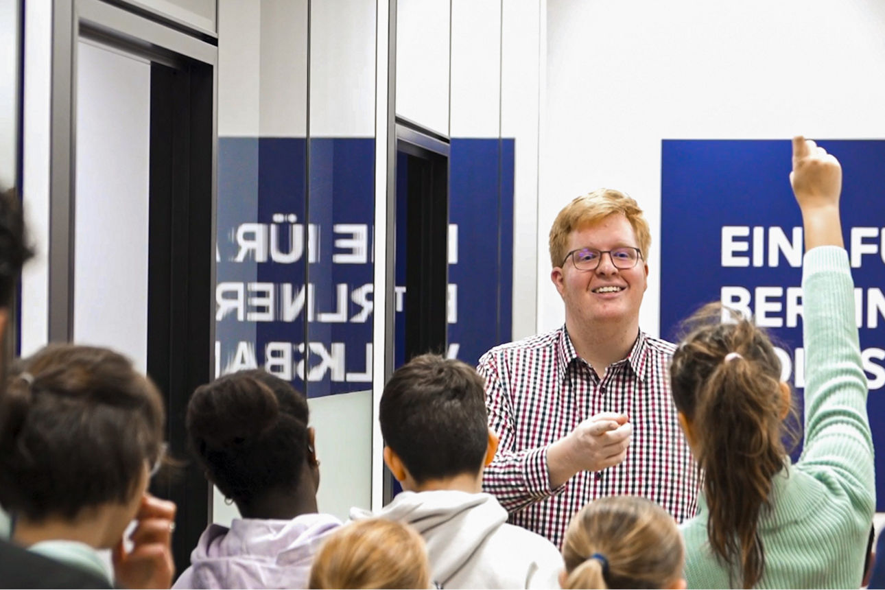 Ein Auszubildender der Berliner Volksbank steht vor Kindern und beantwortet ihre Fragen