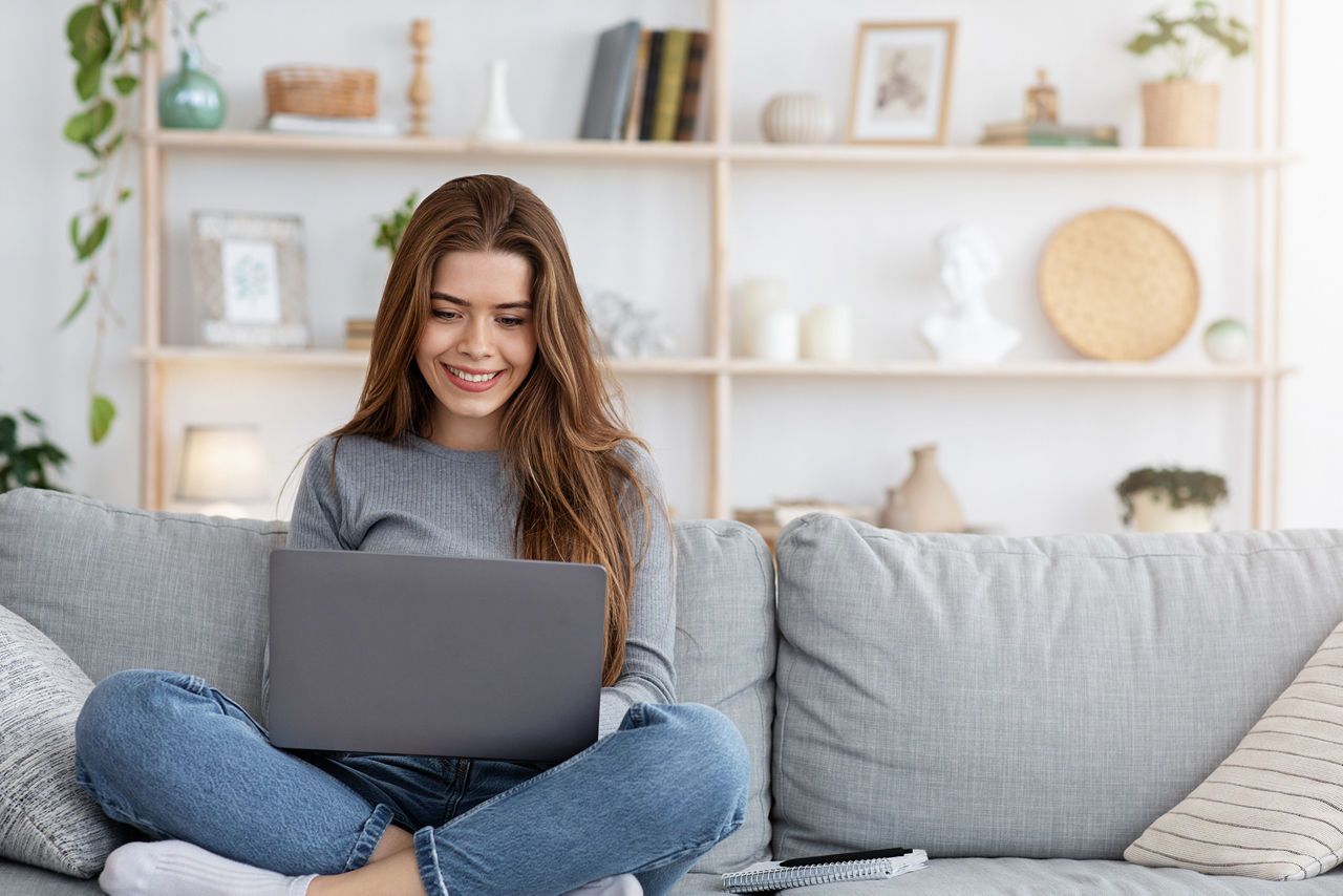 Lächelnde junge Frau, die von zu Hause aus arbeitet, auf der Couch sitzt und Laptop benutzt
