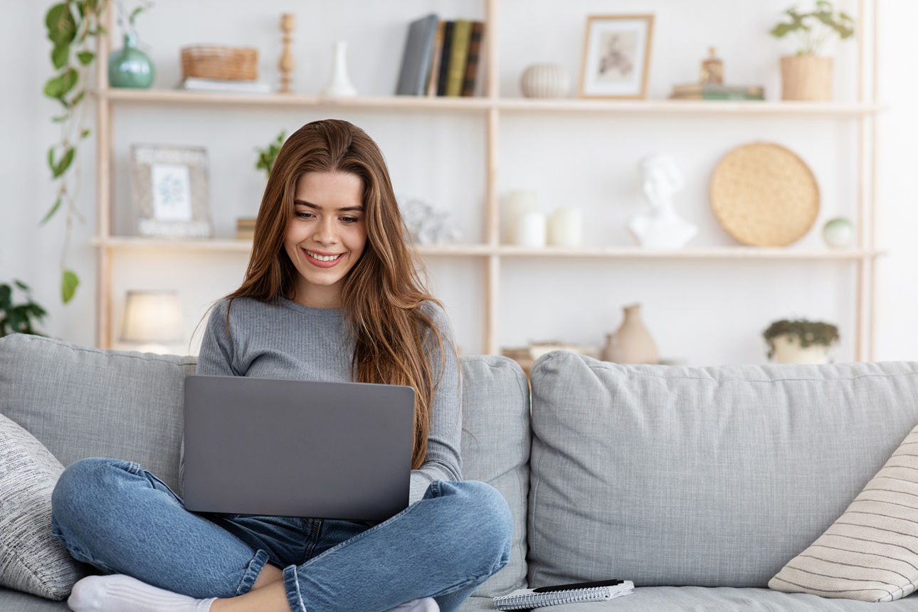 Lächelnde junge Frau, die von zu Hause aus arbeitet, auf der Couch sitzt und Laptop benutzt
