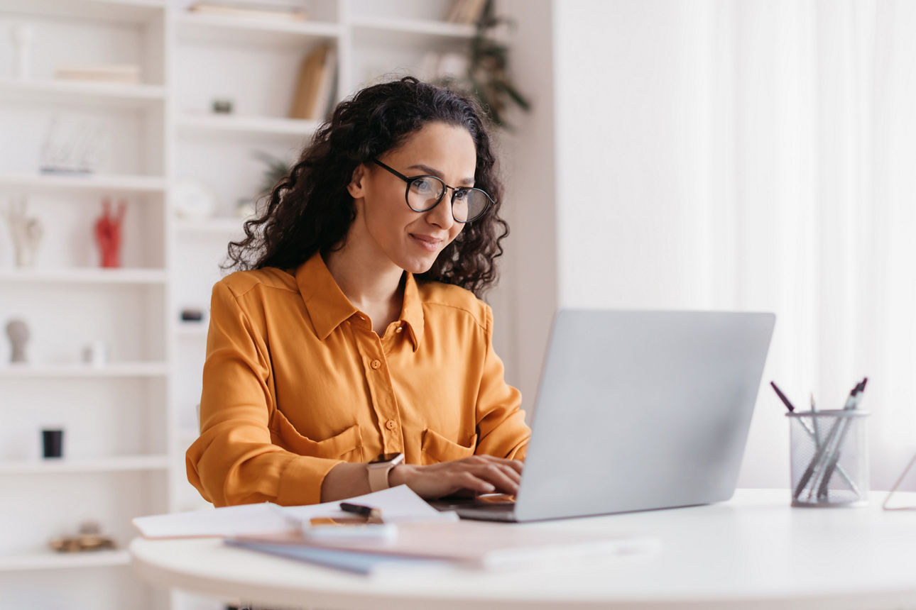 Dame mit Laptop, die online arbeitet und eine Brille trägt, sitzt am Arbeitsplatz im modernen Büro. 