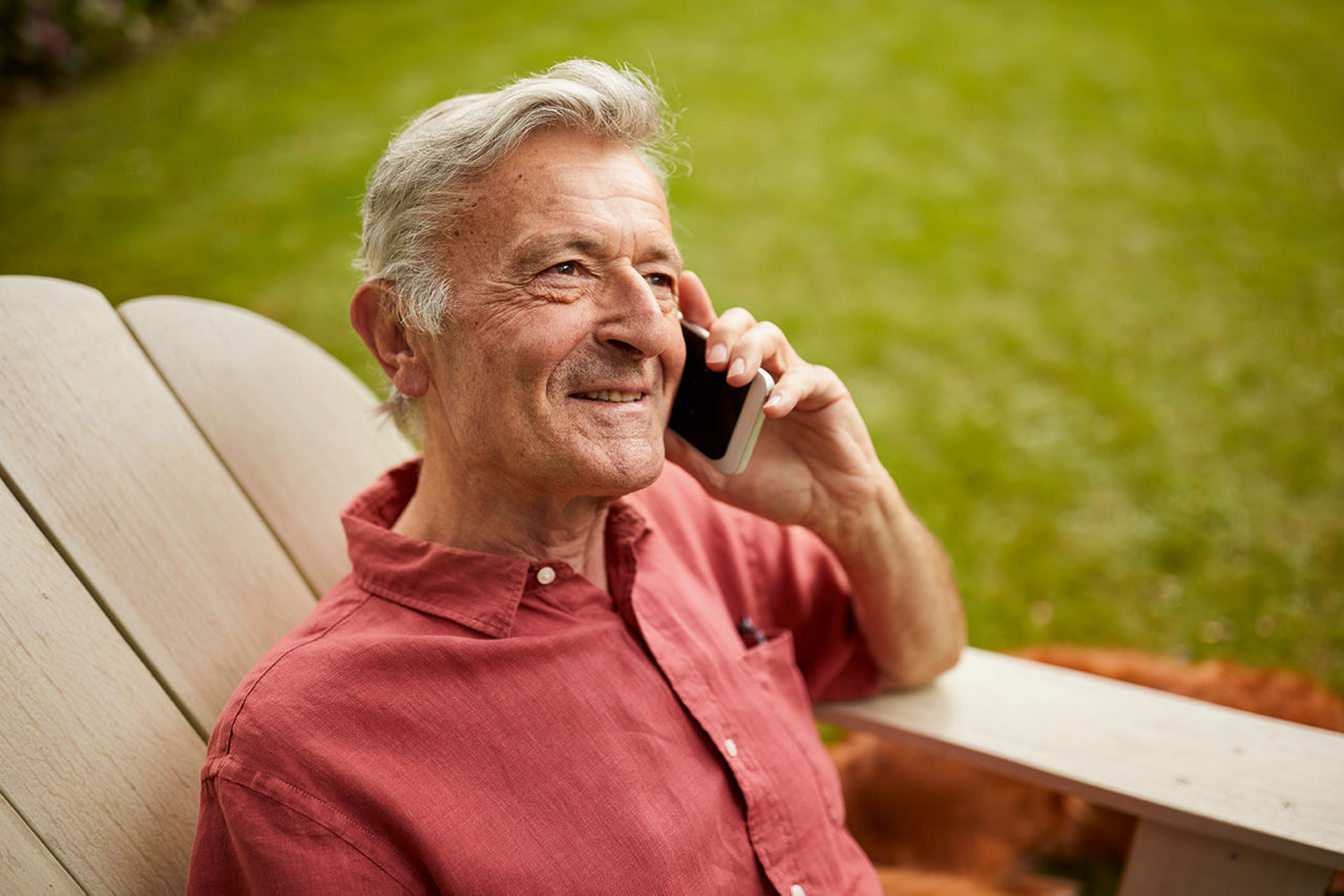 Ältere Person sitzt entspannt im Gartenstuhl und telefoniert mit dem Smartphone – freundlicher Gesichtsausdruck.
