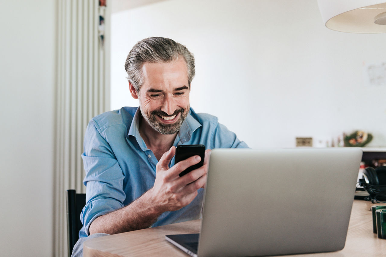 älterer Mann sitzt am Schreibtisch und guckt lächelnd auf sein Handy