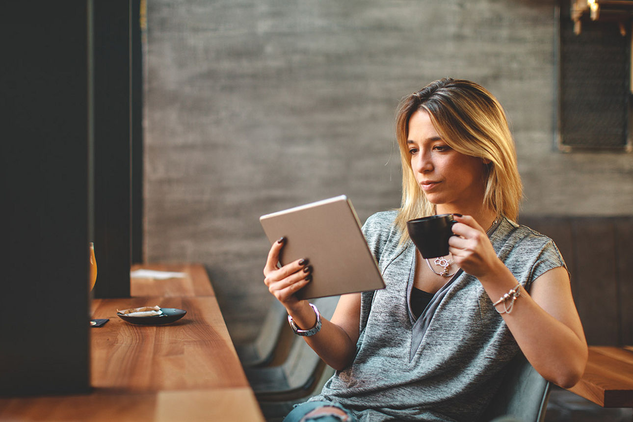 Frau liest mit einer Kaffeetasse in der Hand auf Ihrem Tablet