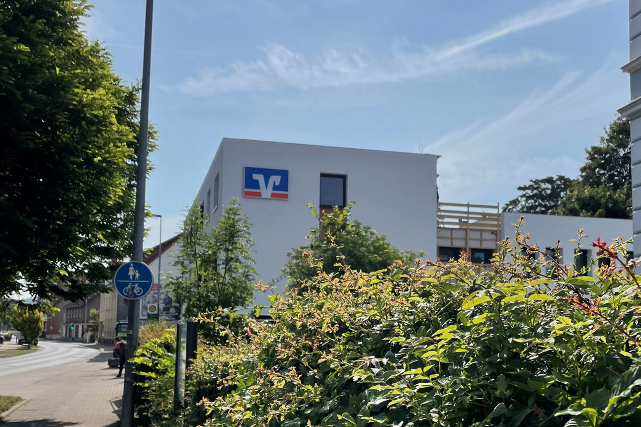 Unsere neue Filiale in Alfeld. Sie sehen das weiße Gebäude mit Flachdach und großen Volksbank Markenzeichen. Davor viele Grünze Pflanzen, abgerundet mit einem blauen Himmel. 