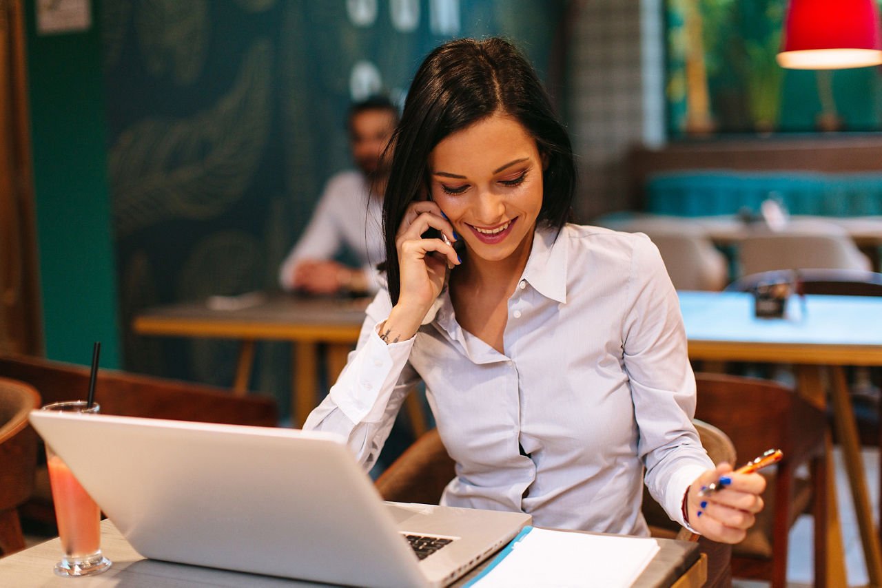 Eine junge Businessfrau sitzt in einem Café vor dem geöffneten Laptop und telefoniert lächelnd