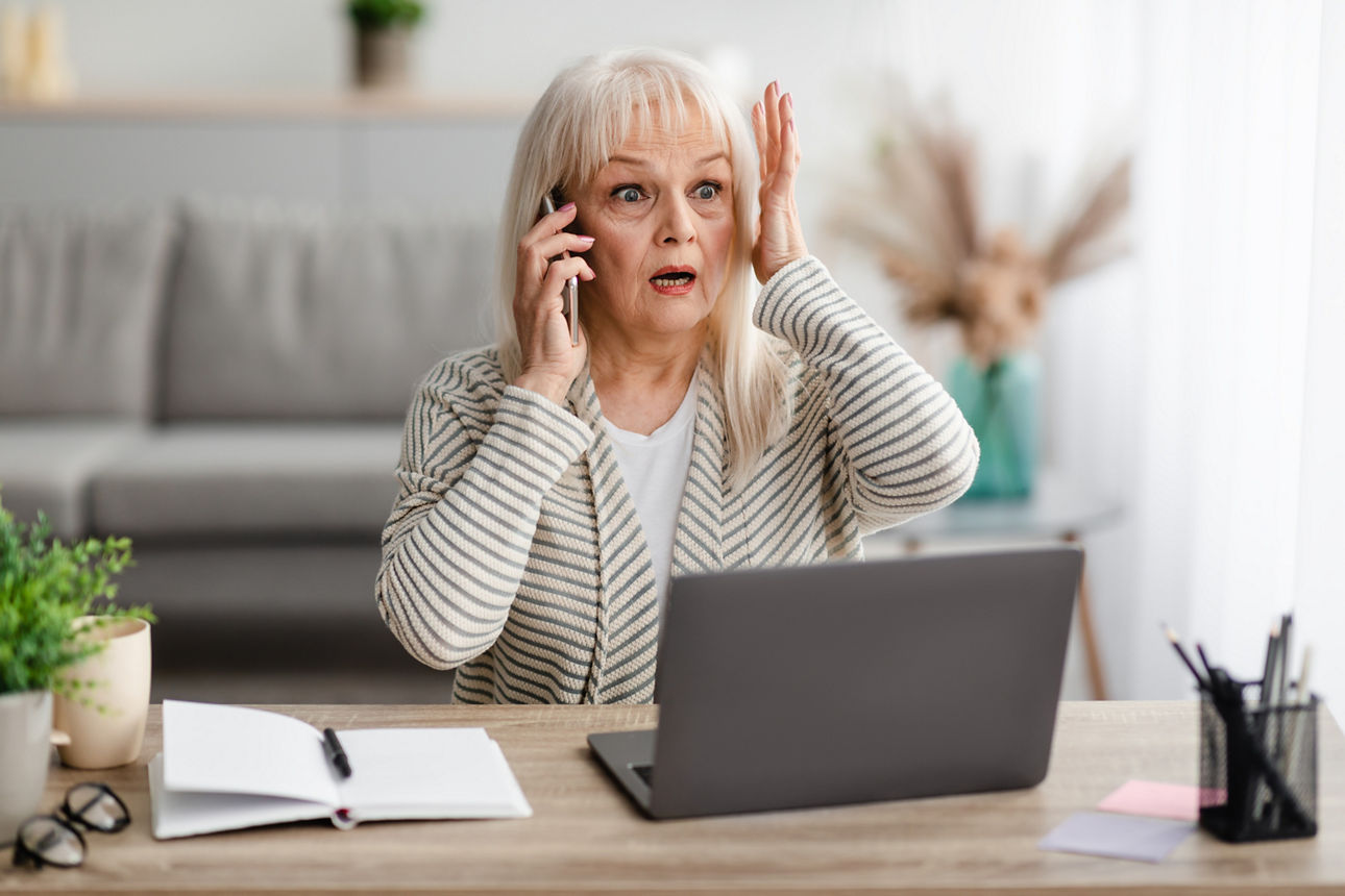 Alte Dame schaut geschockt auf ihren Laptop mit Handy am Ohr