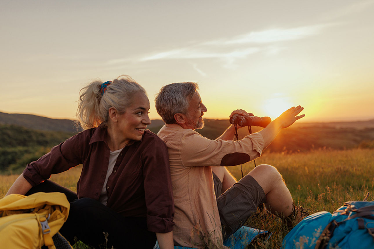 Zwei Personen sitzen bei Sonnenuntergang in einer Wiesenlandschaft und genießen den Ausblick in die Natur.