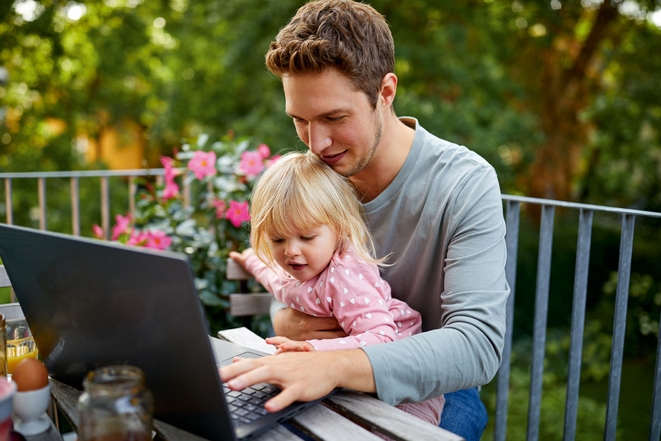 Ein junger Mann sitzt auf dem Balkon und schreibt auf dem Notebook. Seine kleine Tochter sitzt auf seinem Schoß.