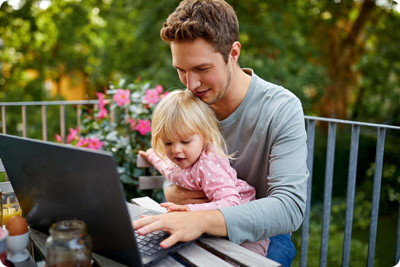 Ein junger Mann sitzt auf dem Balkon und schreibt auf dem Notebook. Seine kleine Tochter sitzt auf seinem Schoß.