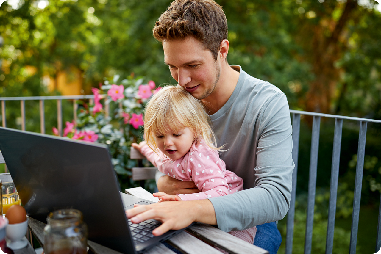 Ein junger Mann sitzt auf dem Balkon und schreibt auf dem Notebook. Seine kleine Tochter sitzt auf seinem Schoß.