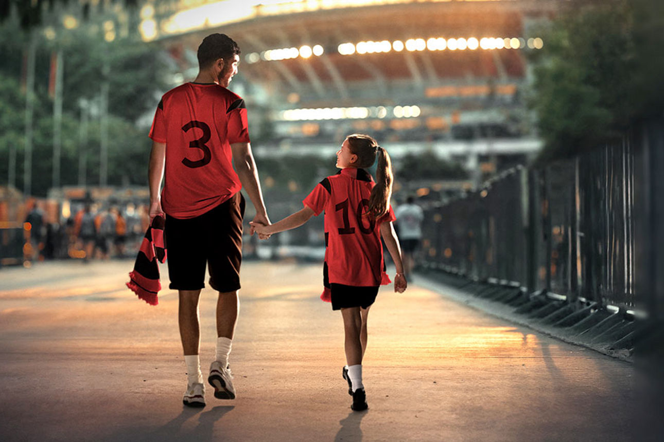 Ein Vater und sein Kind laufen Hand in Hand zum Stadion. Beide tragen Trikots und freuen sich auf das Spiel