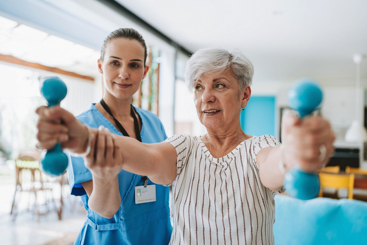 Im Vordergrund sieht man eine ältere Dame. Sie hat weißes Haar und hält in ihren Händen zwei kleine Gewichte. Hinter ihr steht eine junge Therapeutin, die die Frau bei ihrer Physiotherapie unterstützt. 